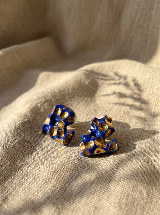 A pair of organic-shaped handmade ceramic stud earrings with deep cobalt blue glaze and shimmering gold accents. Displayed on a beige linen fabric under warm natural sunlight with botanical shadows, highlighting the sculptural texture and handcrafted soul of HECROWN's Jingdezhen artistry.