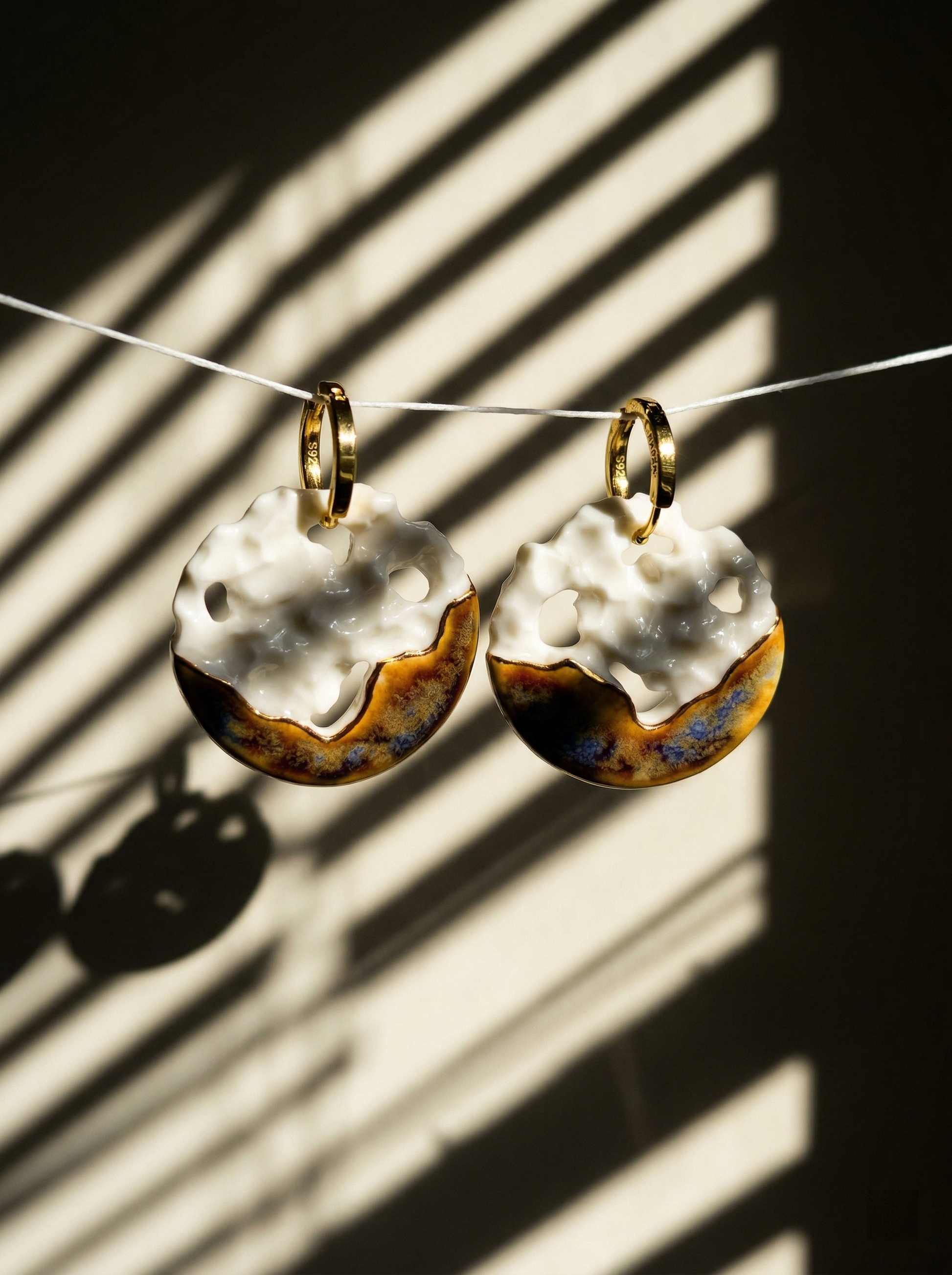 A pair of handmade crater-textured ceramic hoop earrings suspended against a wall, illuminated by warm, slatted window sunlight casting dramatic diagonal shadows. HECROWN artisan jewelry capturing a realistic, moody studio aesthetic that emphasizes the physical texture and sincere craftsmanship of Jingdezhen pottery.