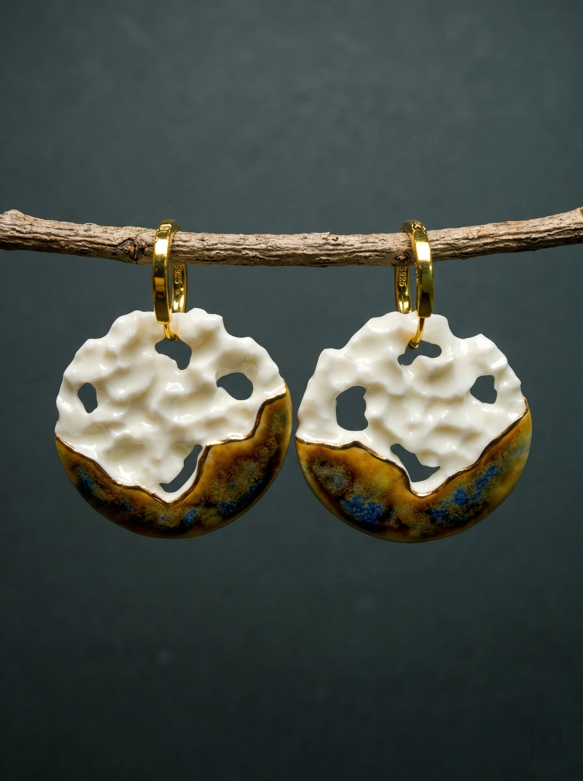 A pair of handmade ceramic hoop earrings with white crater textures and metallic glaze, hanging delicately from a natural wooden branch against a moody dark background. HECROWN artisan jewelry highlighting organic wabi-sabi aesthetics and unique handmade variations in every piece.