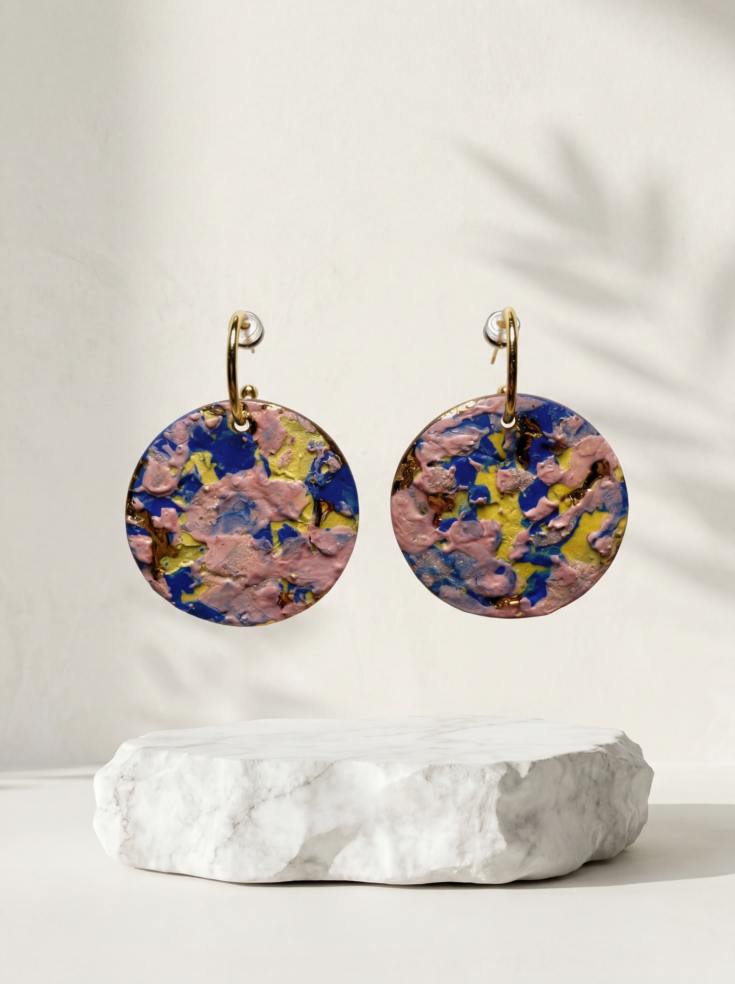 A pair of round handmade ceramic earrings featuring a vibrant, thick impasto glaze in pink, blue, and yellow, suspended above a rough white marble slab. HECROWN contemporary wearable art from Jingdezhen, styled with soft botanical shadows for a bright, modern gallery aesthetic.