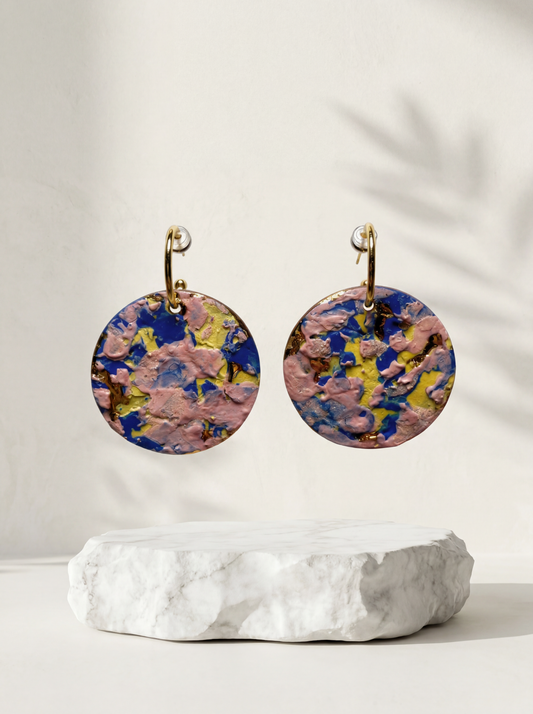 A pair of round handmade ceramic earrings featuring a vibrant, thick impasto glaze in pink, blue, and yellow, suspended above a rough white marble slab. HECROWN contemporary wearable art from Jingdezhen, styled with soft botanical shadows for a bright, modern gallery aesthetic.