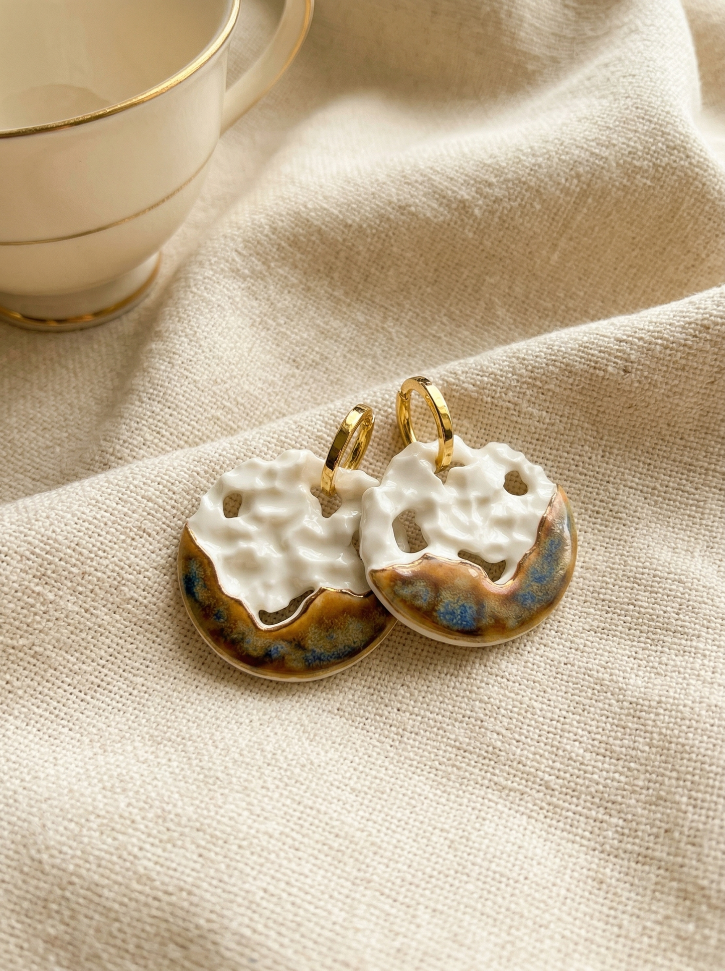 Close-up of 1-of-1 handmade ceramic hoop earrings featuring an uneven white crater-like texture contrasting with a metallic brown and blue glaze. HECROWN signature artisan jewelry resting on unbleached linen next to a vintage teacup, capturing a quiet luxury and wabi-sabi aesthetic.