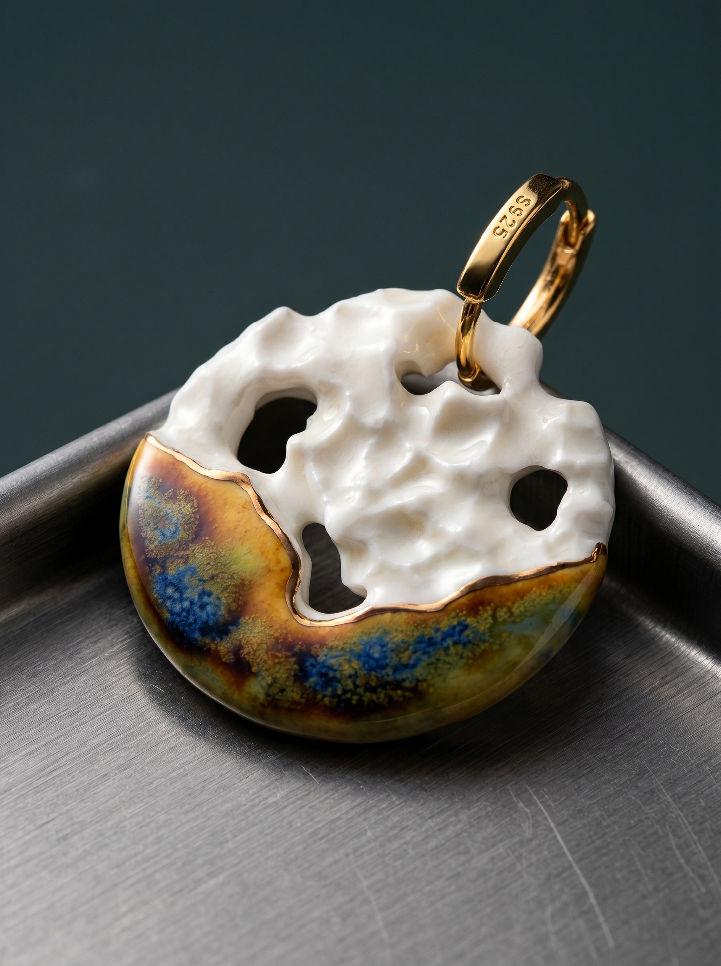 Close-up of a single HECROWN handmade ceramic earring featuring white crater textures and a gold S925 hoop, resting on a cold, brushed steel tray. Showcasing a striking aesthetic contrast between organic earthy pottery and sharp industrial metal.