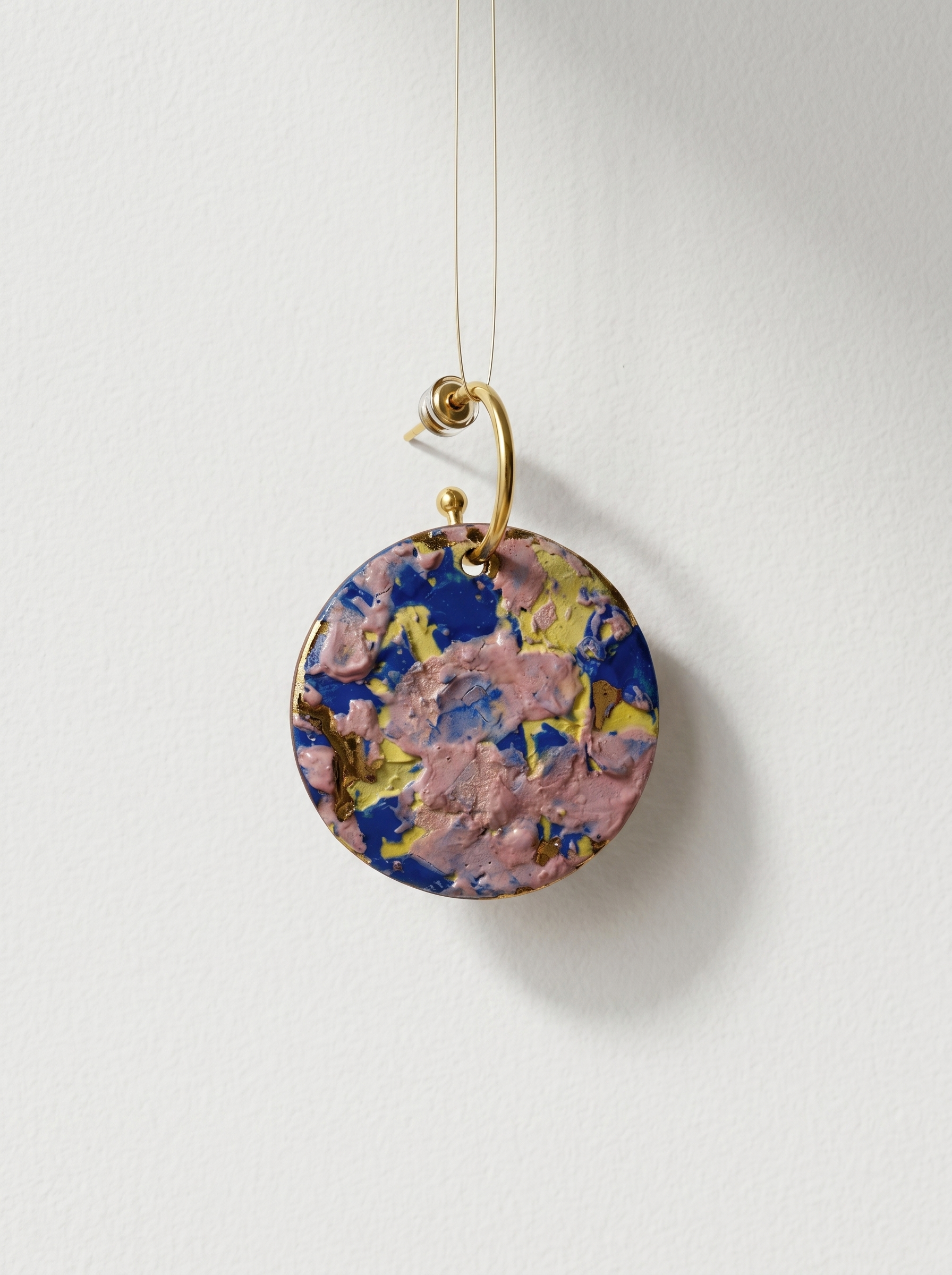 A single handmade ceramic earring with vibrant pink, blue, and yellow impasto glaze, suspended on a thin wire against a clean white wall. HECROWN 1-of-1 wearable art collection from Jingdezhen, highlighting the sculptural design and lightweight floating aesthetic.