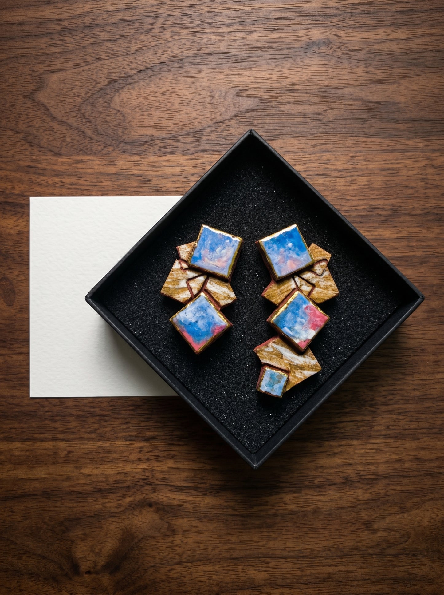 Top-down view of HECROWN geometric ceramic earrings in a premium black jewelry box, accompanied by a white artisan thank-you card on a walnut wood surface. Showcasing the sincere unboxing experience and handcrafted details from Jingdezhen.