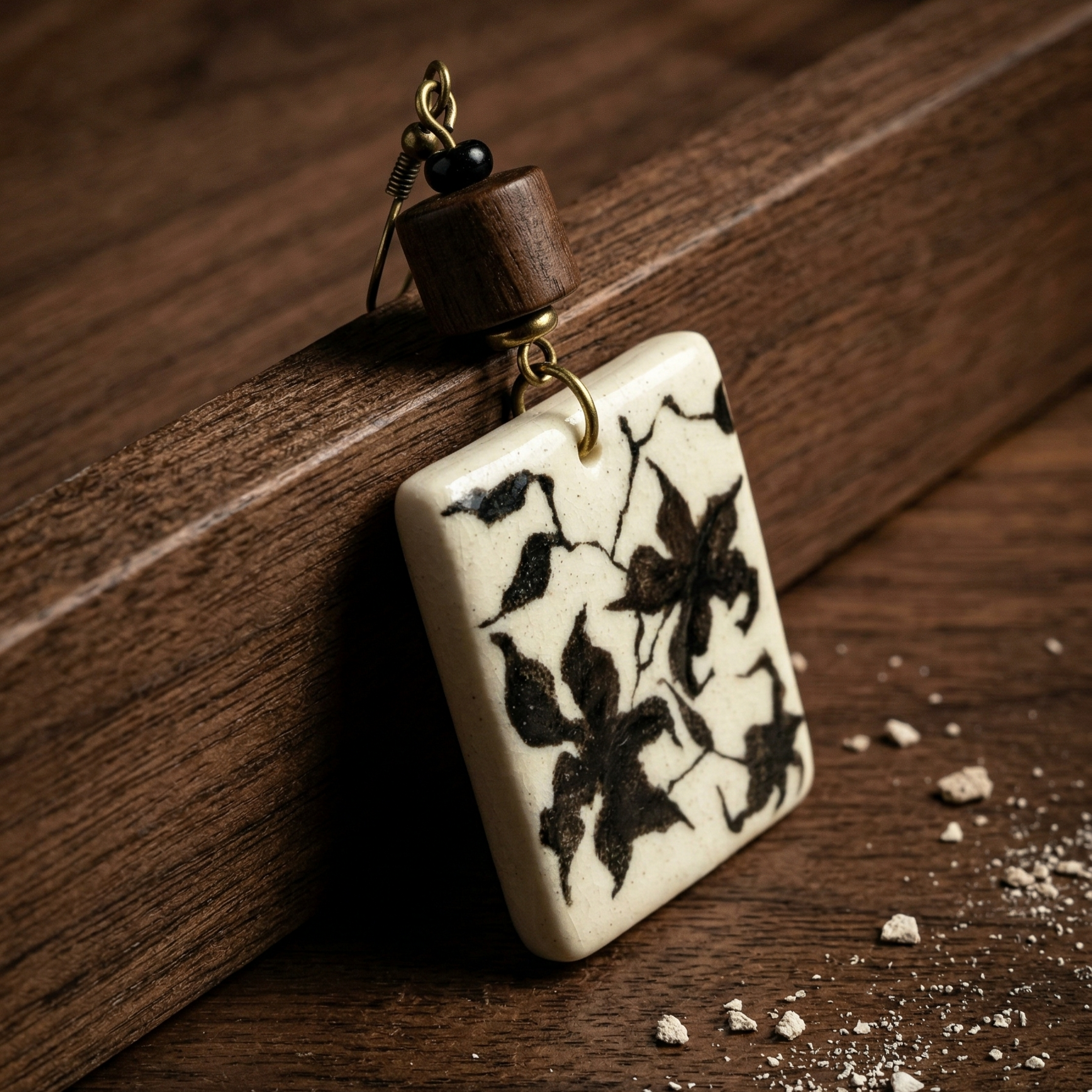 Close-up of a wabi-sabi rectangular ceramic earring featuring dark botanical ink motifs, antiqued brass hardware, and a wooden bead, resting on textured dark walnut wood. HECROWN artisan jewelry handcrafted in Jingdezhen, conveying a vintage, earthy, and quiet luxury aesthetic.