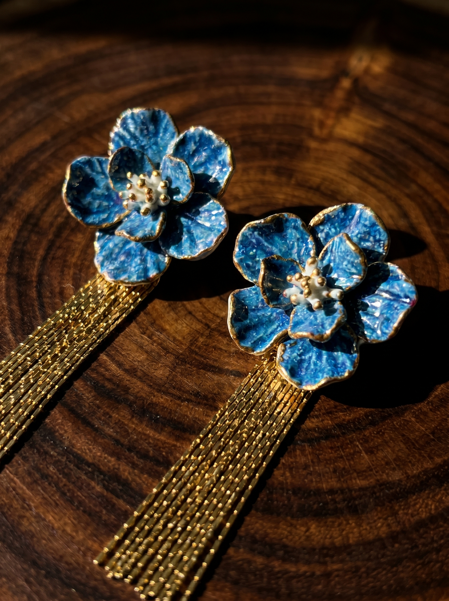 Close-up of HECROWN blue porcelain flower earrings with long gold tassels on a dark wood grain surface. Showcasing the sincere handcrafted texture and vibrant glaze from Jingdezhen.