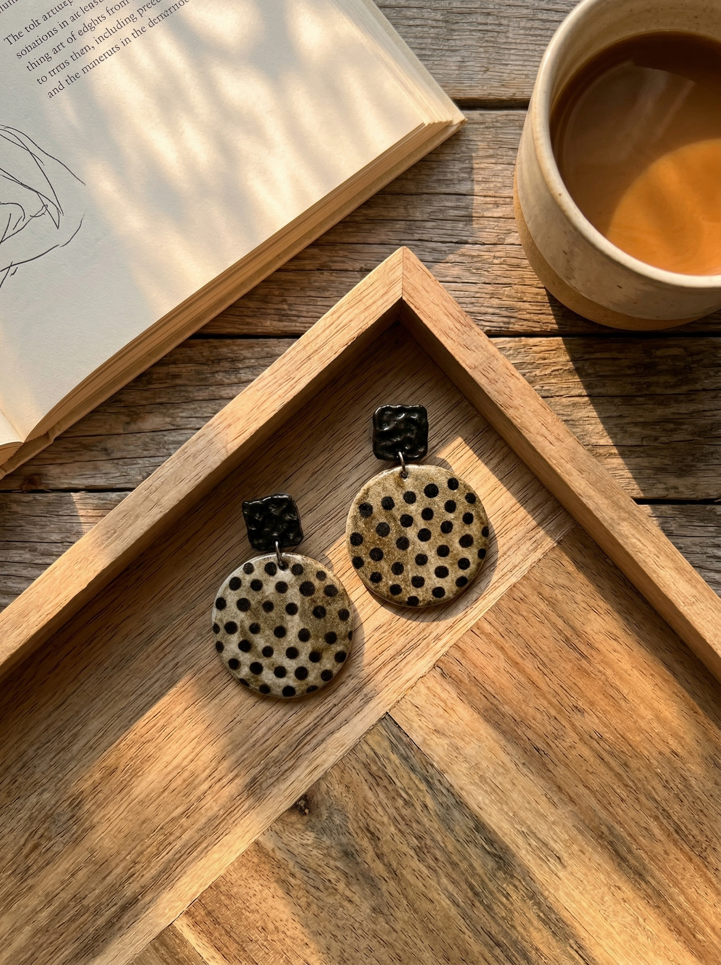 HECROWN round speckled ceramic earrings with black hammered tops resting on a wooden tray next to a coffee cup. A warm lifestyle shot of Jingdezhen handmade jewelry.