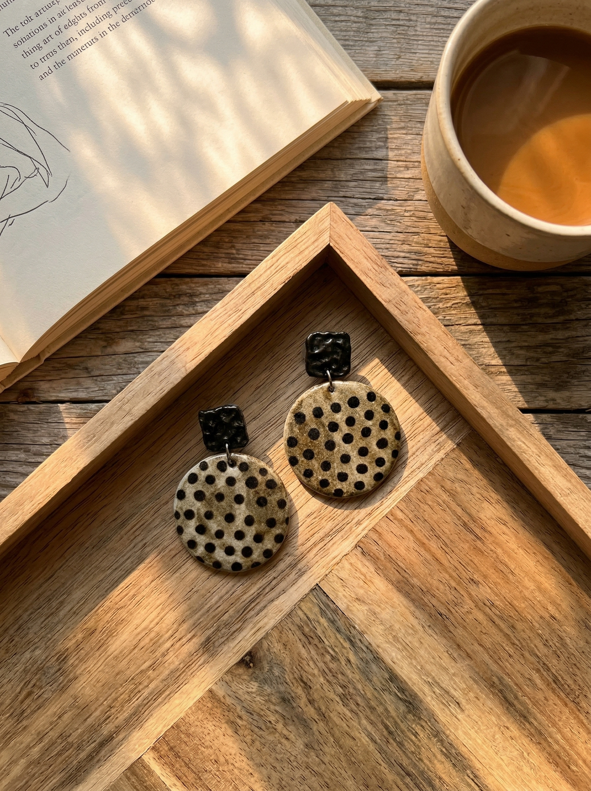 HECROWN round speckled ceramic earrings with black hammered tops resting on a wooden tray next to a coffee cup. A warm lifestyle shot of Jingdezhen handmade jewelry.