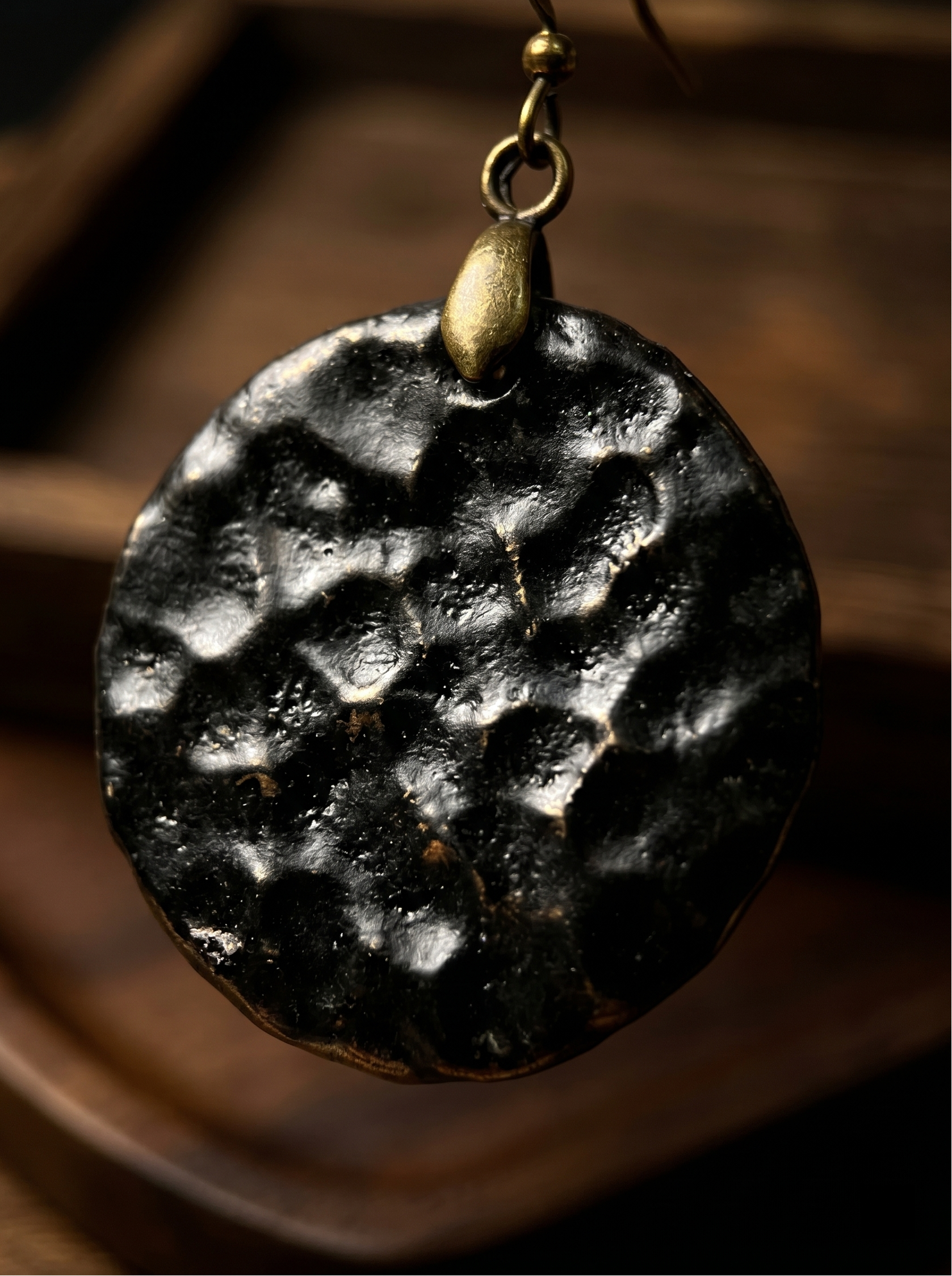 Extreme macro close-up of a HECROWN black ceramic earring, highlighting the deep hammered crater texture and vintage brass hook. Sincere artisan craftsmanship.