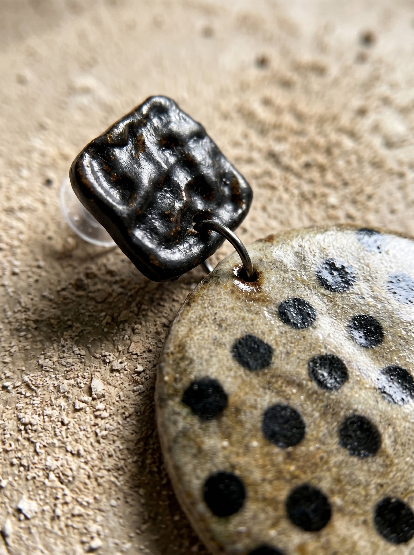 Macro close-up of a HECROWN ceramic earring on rough stone, highlighting the hammered black top and beige speckled polka dot texture. Authentic handcrafted artisan jewelry details.
