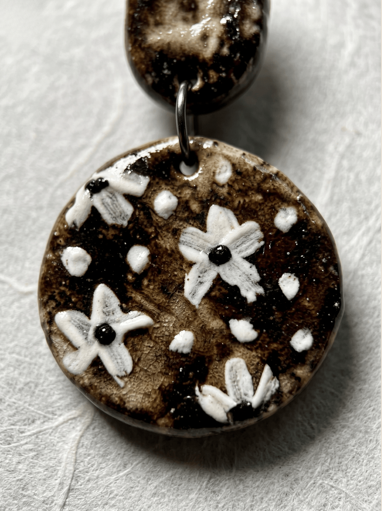 Extreme macro shot of a HECROWN ceramic earring highlighting the raised impasto texture of white painted flowers on a dark crackle glaze. Authentic Jingdezhen craftsmanship.