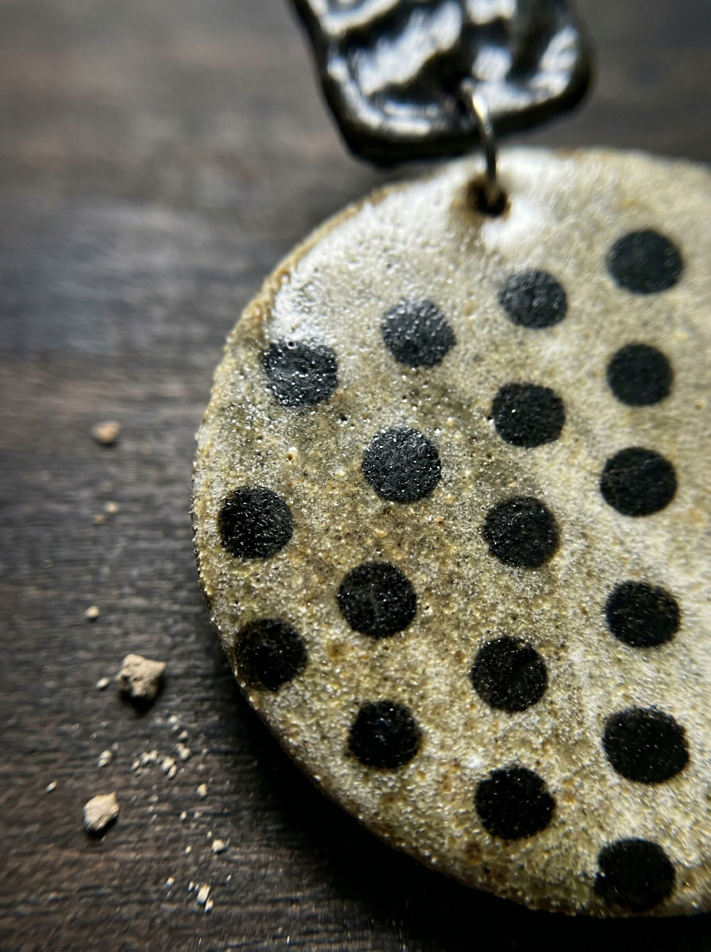 Extreme macro shot of HECROWN round speckled ceramic earring on dark wood with clay dust. Highlighting the raw, sincere wabi-sabi texture of genuine handcrafted artisan jewelry.