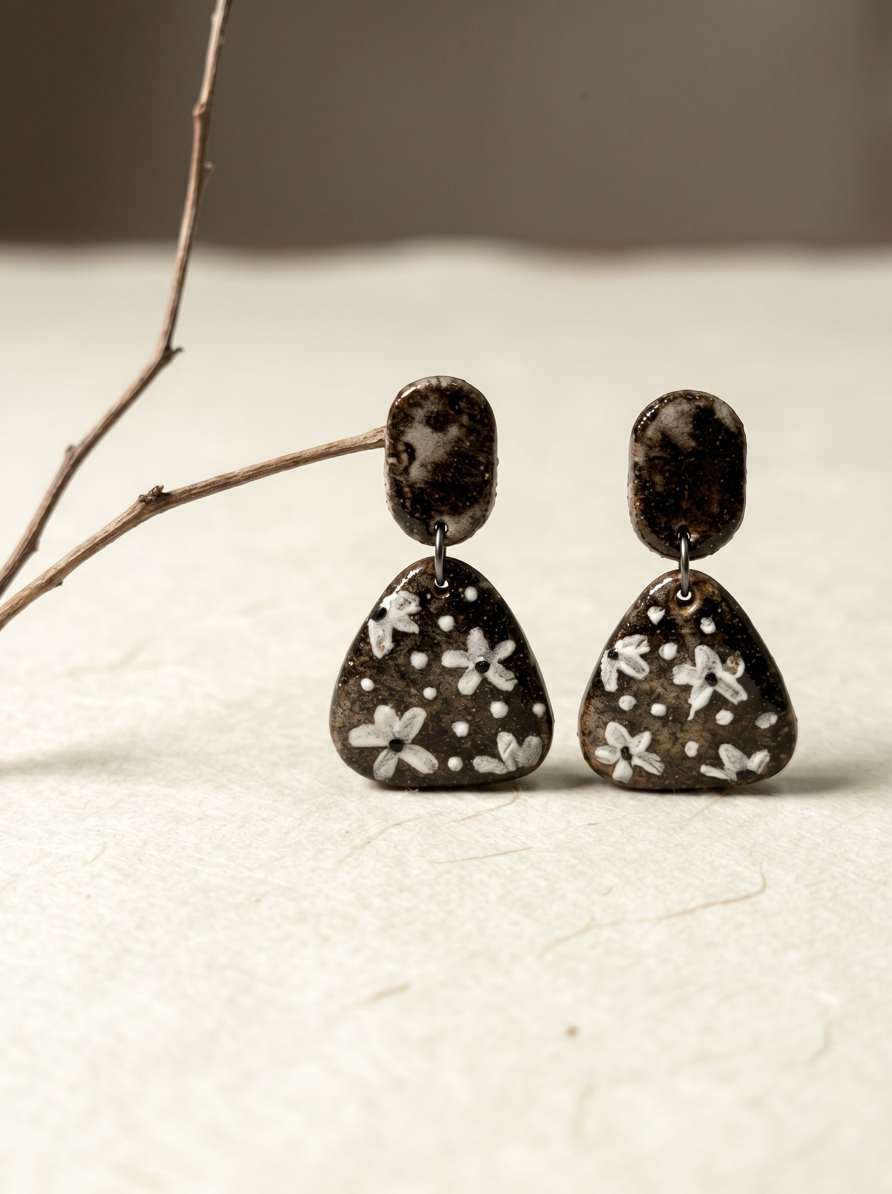 Close-up of a HECROWN ceramic earring on rough stone, highlighting the hammered black top and beige speckled polka dot texture. Authentic handcrafted artisan jewelry details.