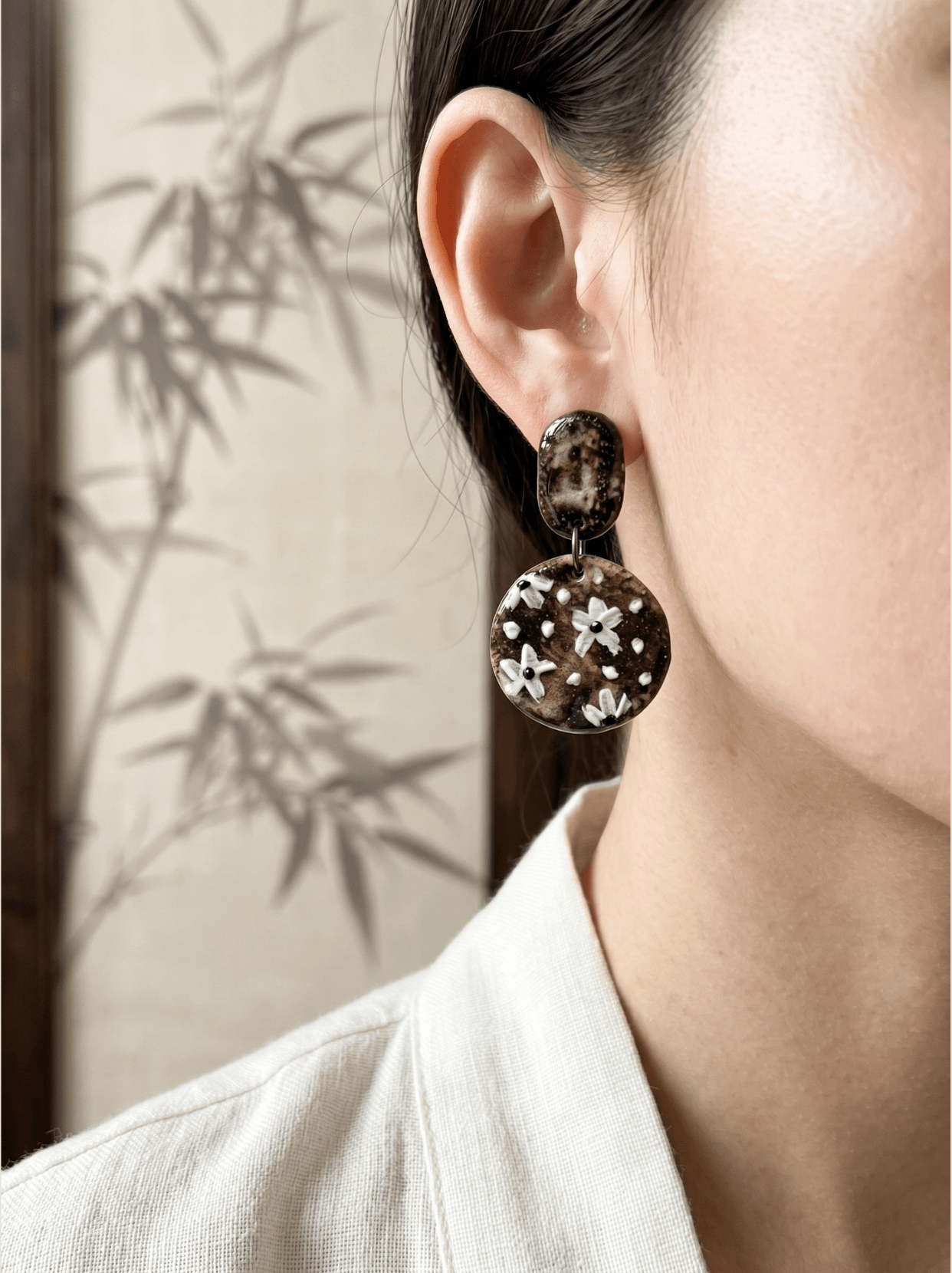 Side profile of a model wearing HECROWN round hand-painted floral ceramic earrings against a bamboo background. Showcasing elegant, everyday artisan handmade jewelry.
