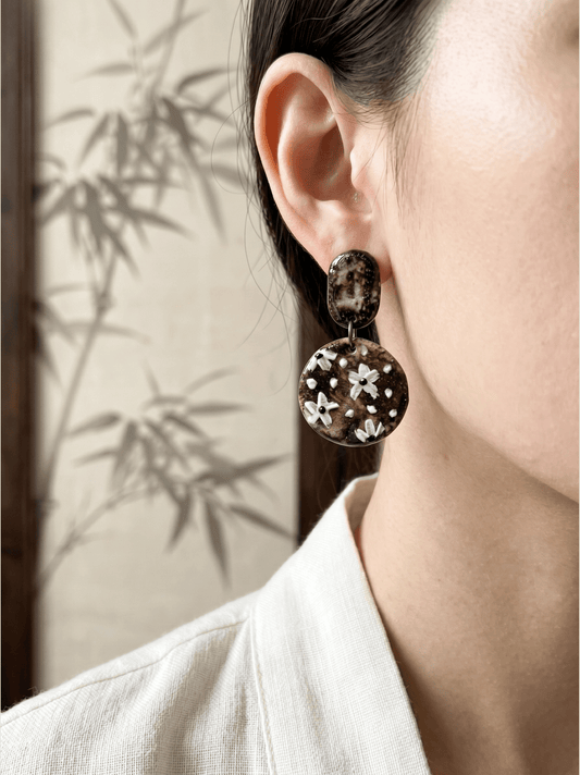 Side profile of a model wearing HECROWN round hand-painted floral ceramic earrings against a bamboo background. Showcasing elegant, everyday artisan handmade jewelry.