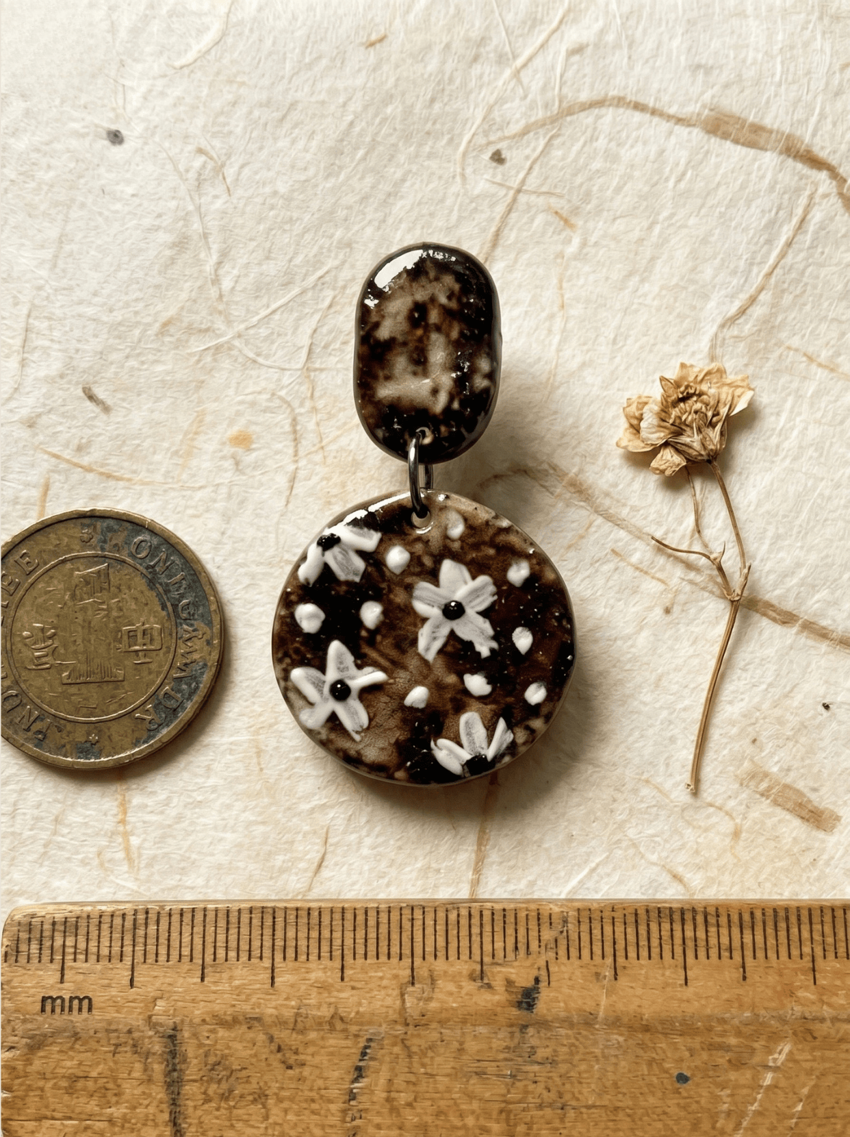 Size reference for HECROWN round painted floral ceramic earring, displayed with a wooden mm ruler, a coin, and a dried flower. Jingdezhen handmade jewelry scale.