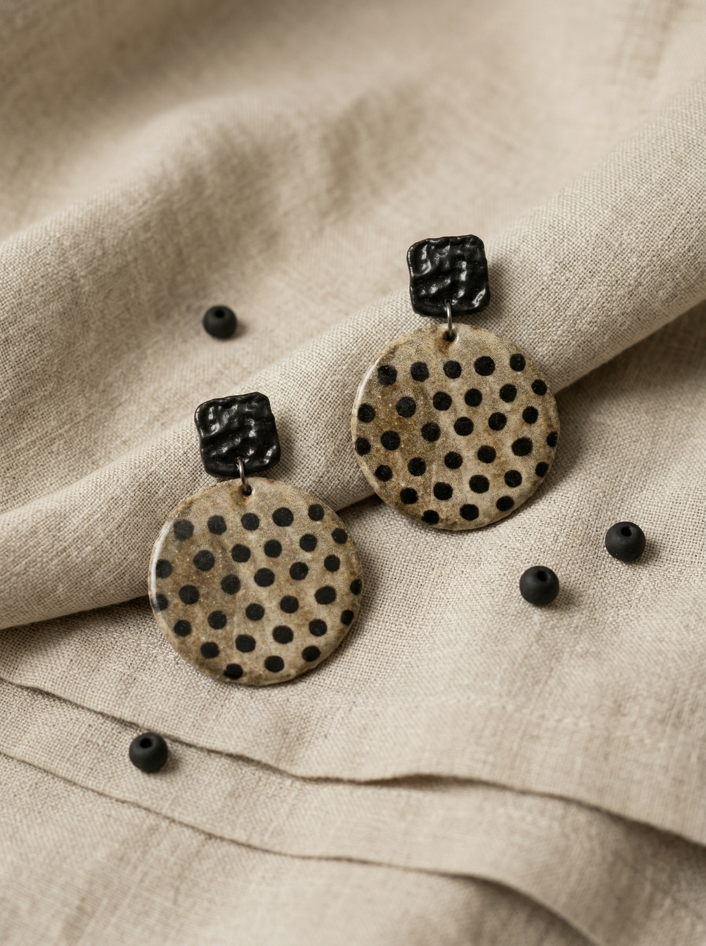 A pair of HECROWN round speckled ceramic earrings with black polka dots on textured linen. Authentic Jingdezhen handmade jewelry perfect for everyday artisan wear.