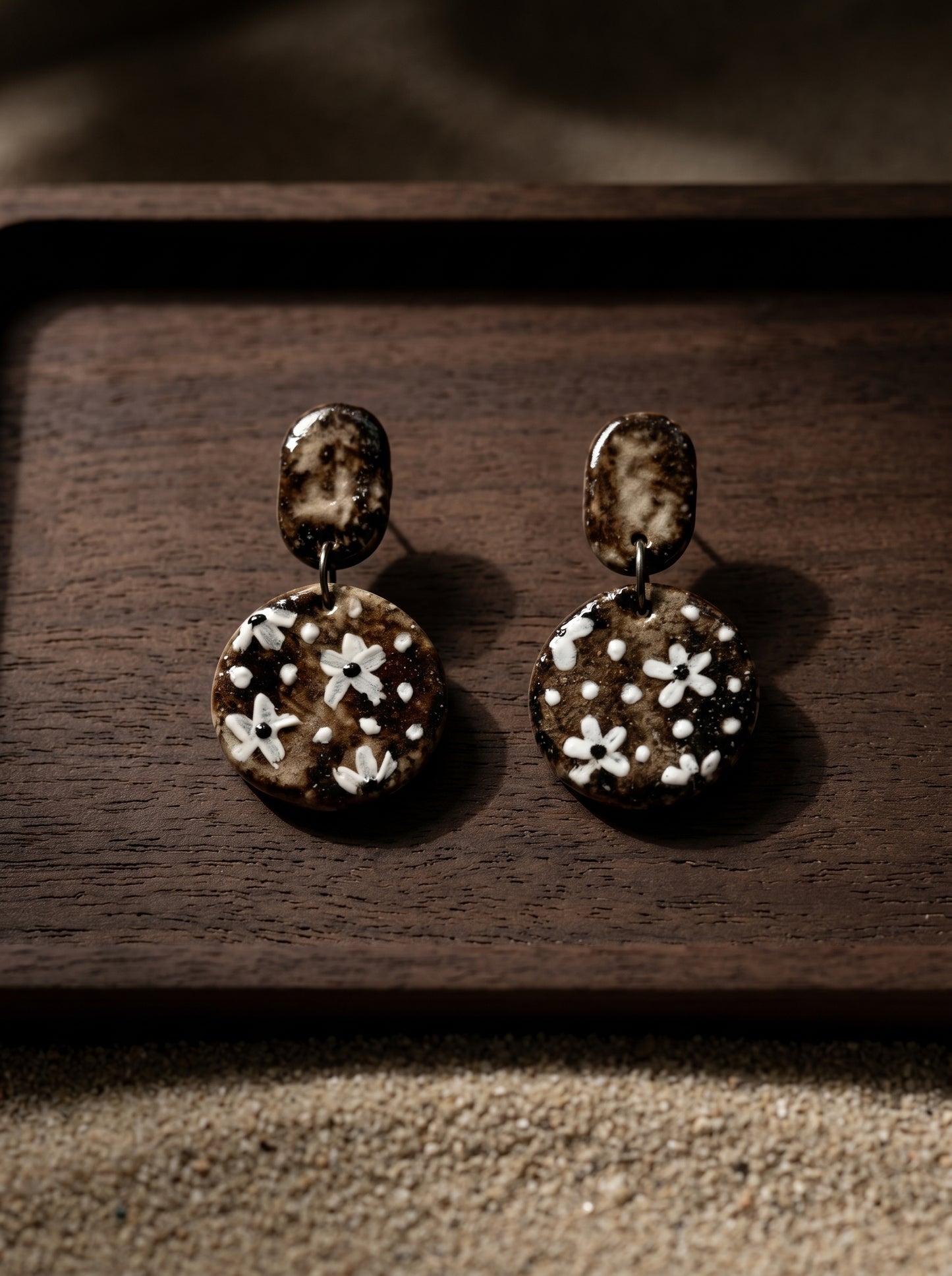 A pair of HECROWN round vintage ceramic earrings with hand-painted white flowers on a dark wood tray. Sincere Jingdezhen handmade jewelry and a unique anniversary gift.