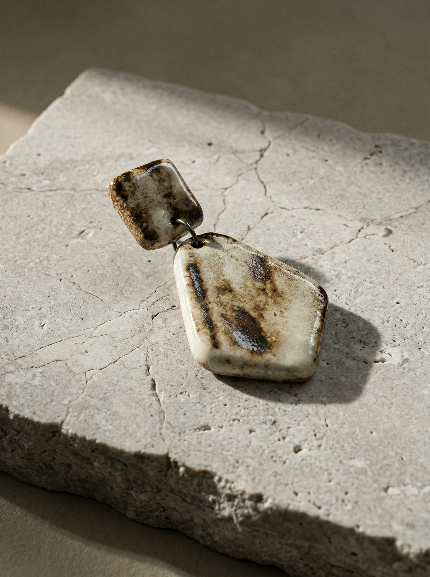 A single HECROWN geometric ceramic earring resting on a textured stone slab in sunlight. The earthy brown and white glaze highlights the sincere craftsmanship of Jingdezhen handmade jewelry, perfect for an anniversary gift.