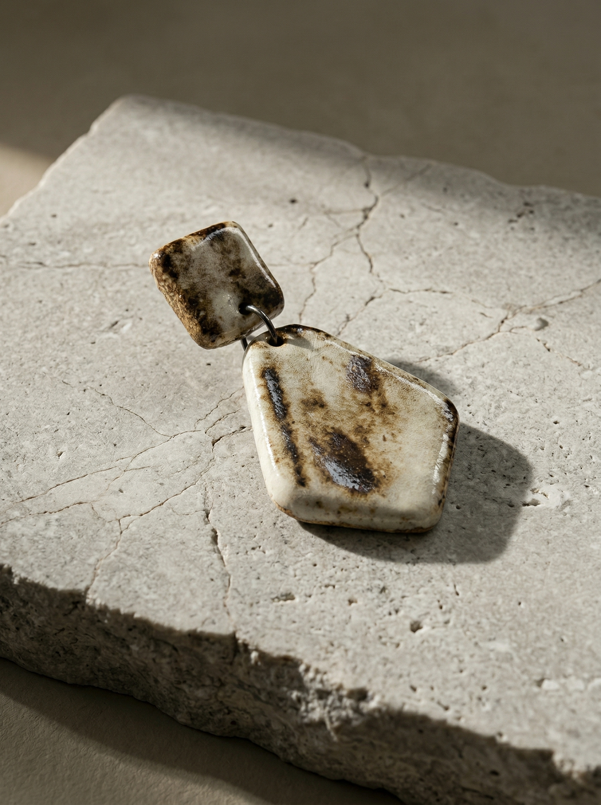 A single HECROWN geometric ceramic earring resting on a textured stone slab in sunlight. The earthy brown and white glaze highlights the sincere craftsmanship of Jingdezhen handmade jewelry, perfect for an anniversary gift.