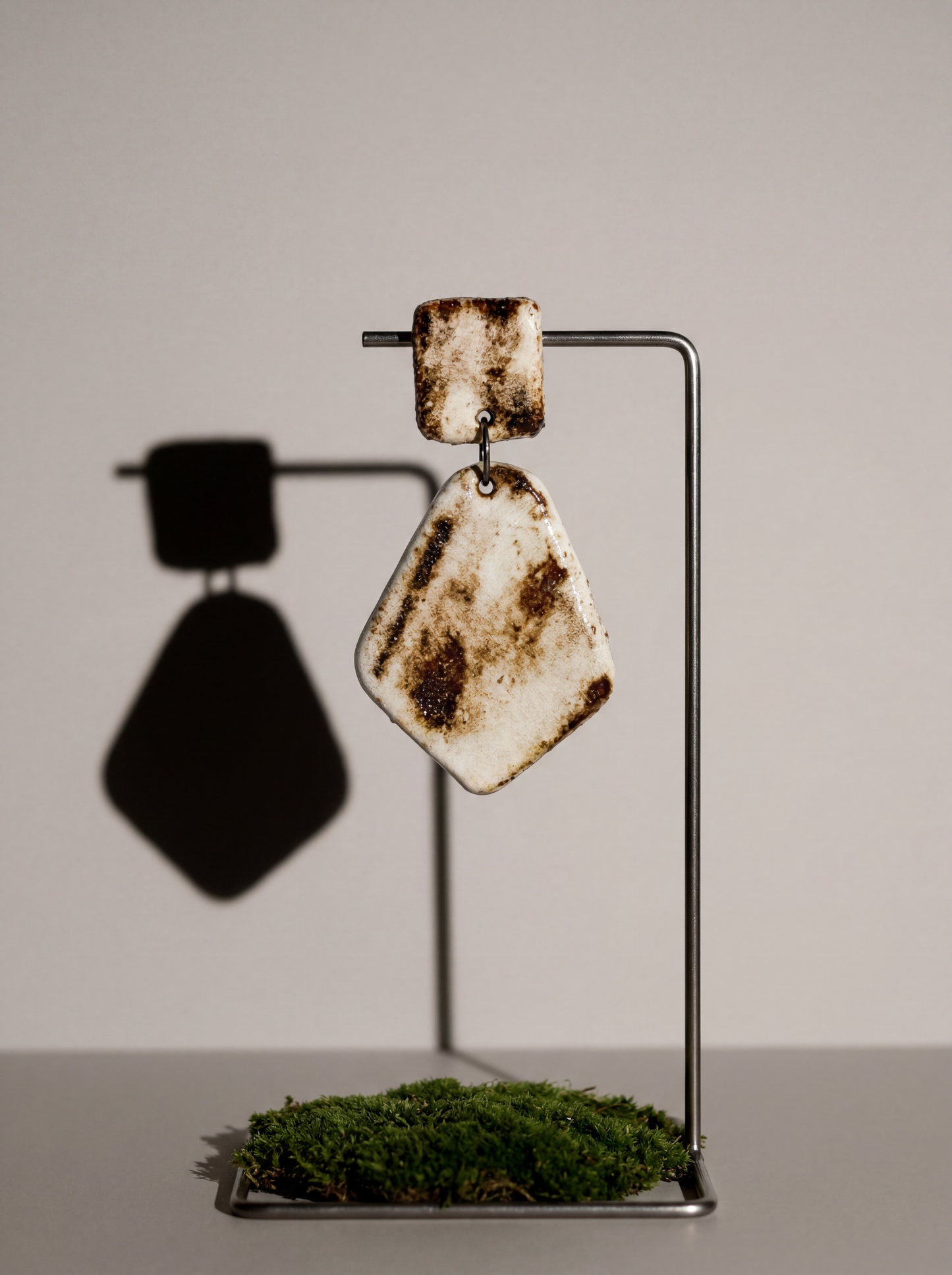 A HECROWN rustic ceramic earring suspended on a metal stand above green moss. Blending earthy kiln-fired textures with modern design, an ideal Valentine's Day gift for lovers of handmade jewelry.