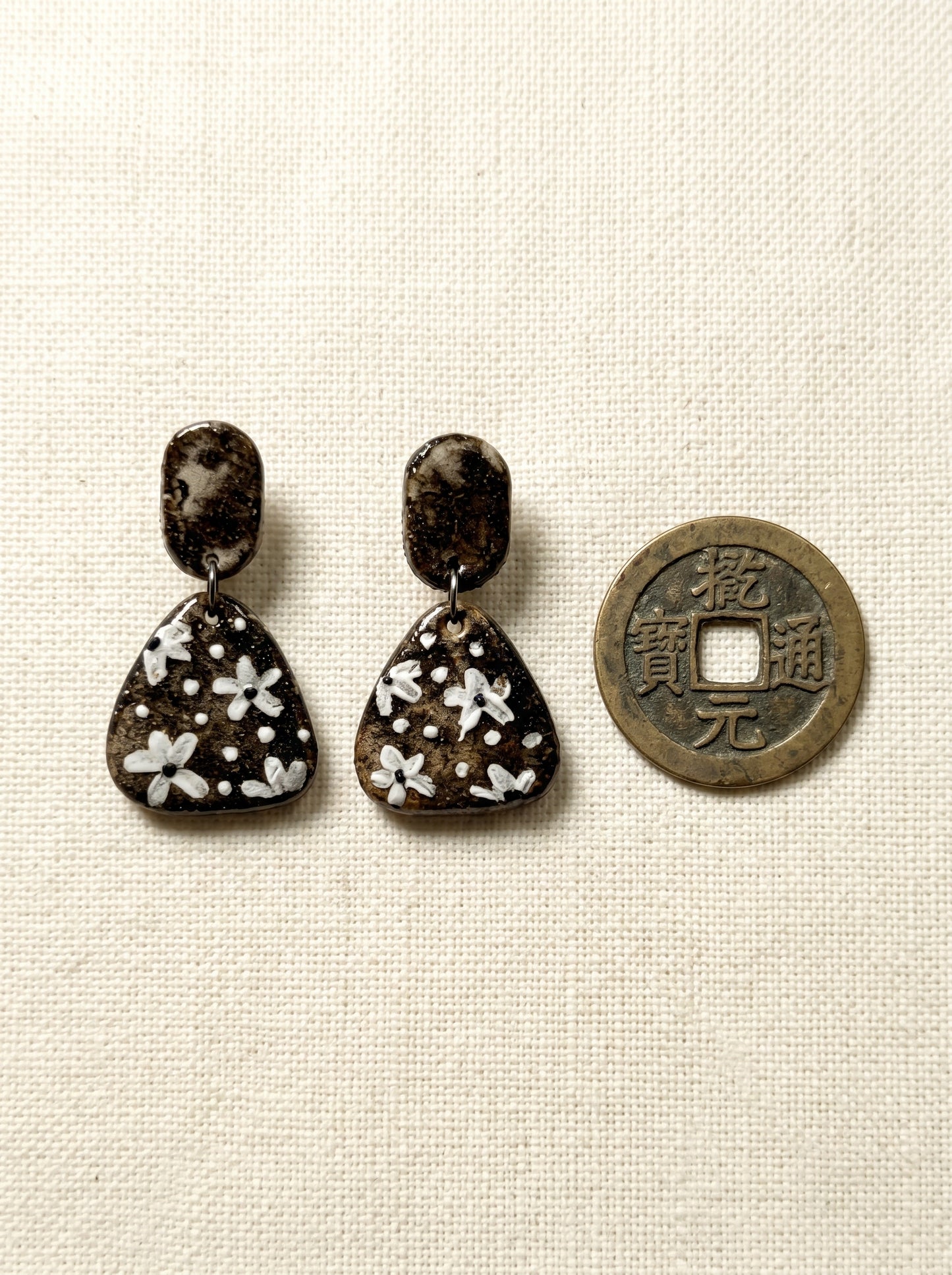 A pair of HECROWN vintage ceramic earrings with hand-painted white flowers on a dark textured glaze, displayed next to an antique coin. Authentic Jingdezhen handmade jewelry.