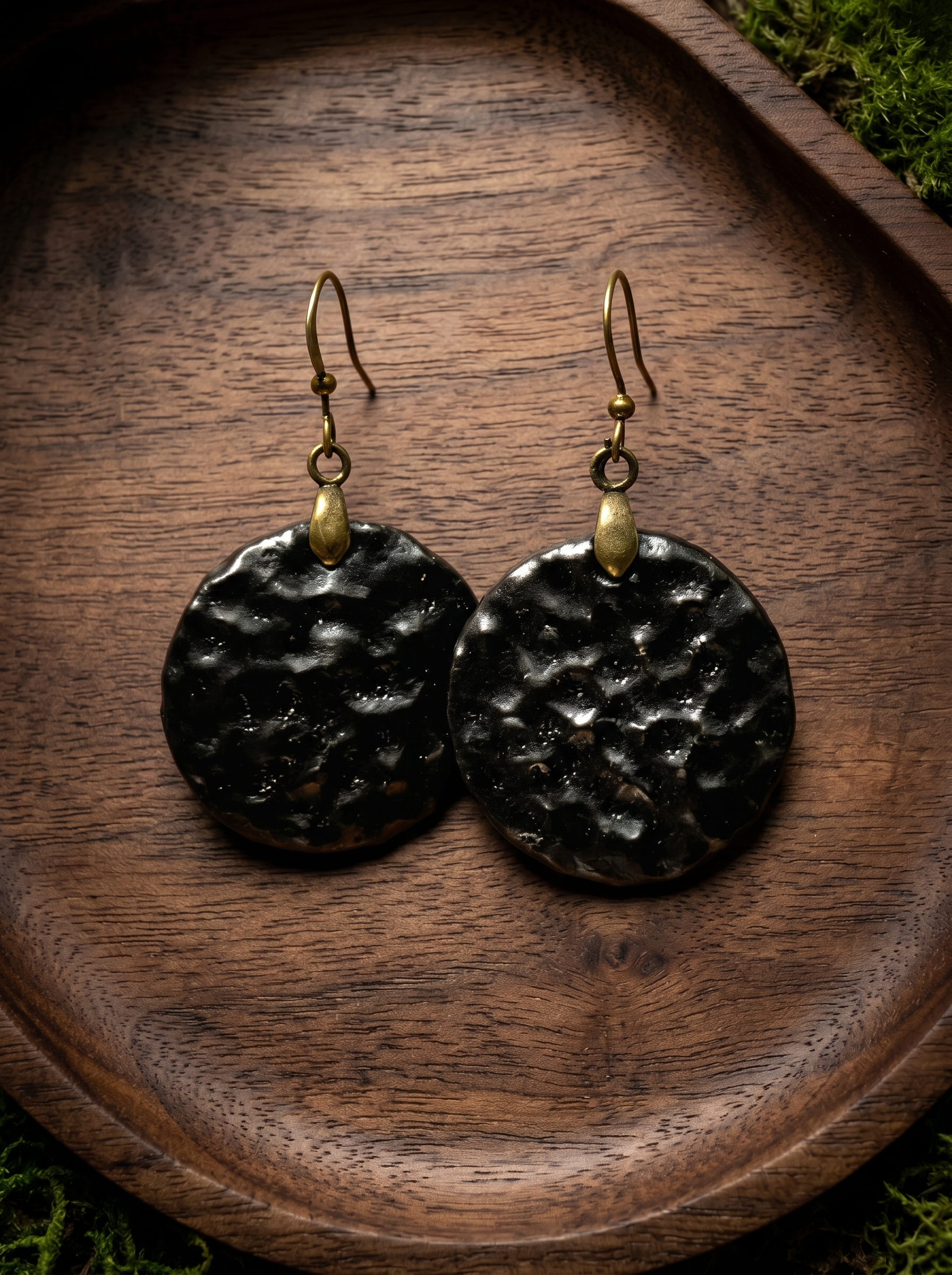 A pair of HECROWN round black ceramic earrings with a hammered crater texture, resting on a dark wooden tray. Authentic Jingdezhen handmade jewelry.