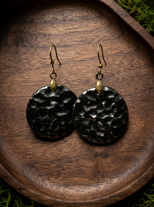 A pair of HECROWN round black ceramic earrings with a hammered crater texture, resting on a dark wooden tray. Authentic Jingdezhen handmade jewelry.