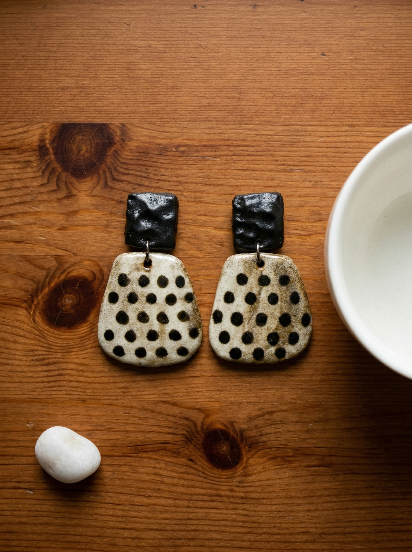 A pair of HECROWN chunky black and white speckled ceramic statement earrings resting on a wooden table. A unique wabi-sabi artisan anniversary gift from Jingdezhen.