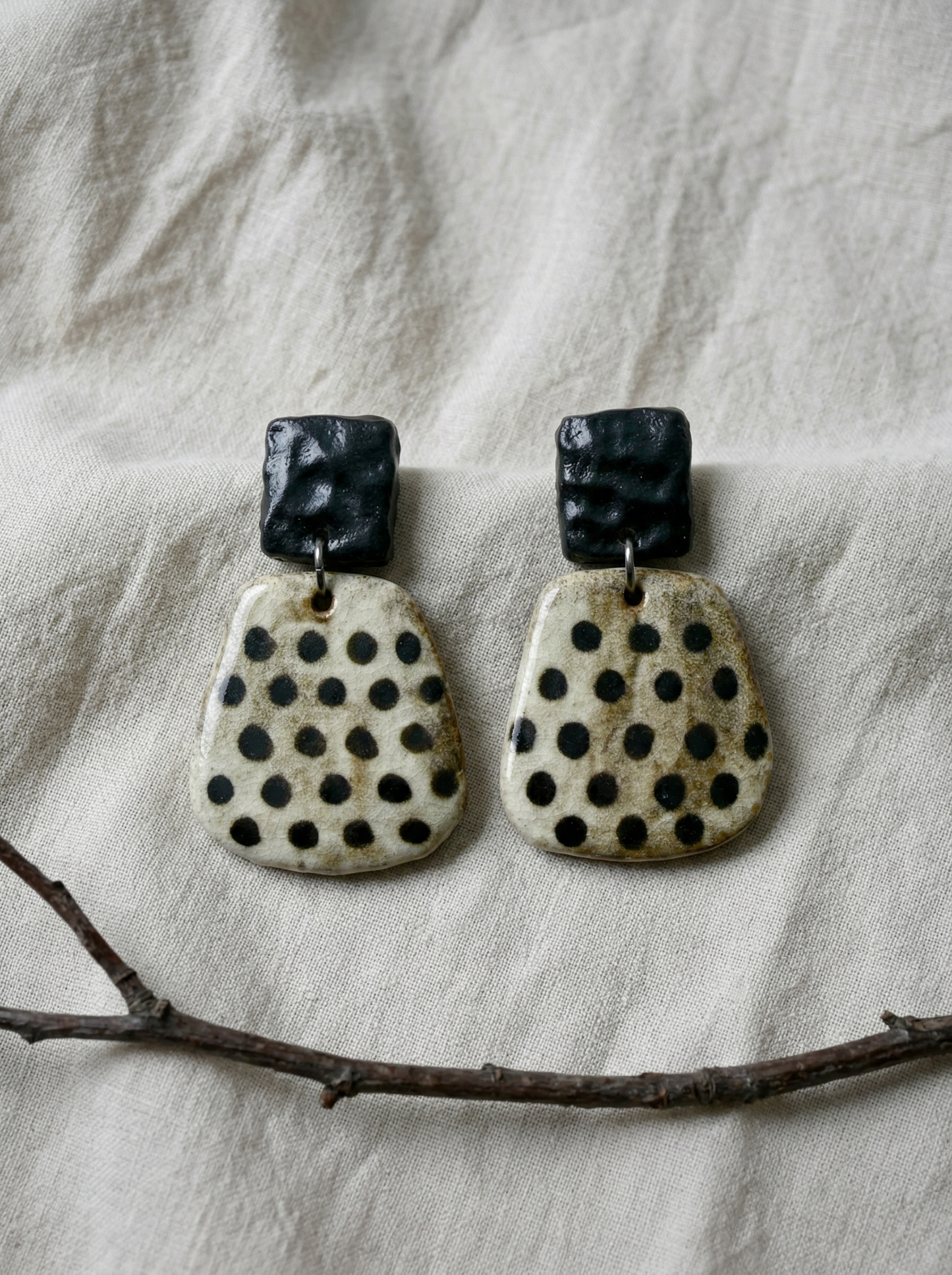 A pair of HECROWN wabi-sabi ceramic earrings with black hammered tops and speckled dot bottoms on natural linen. Unique handmade jewelry from Jingdezhen.