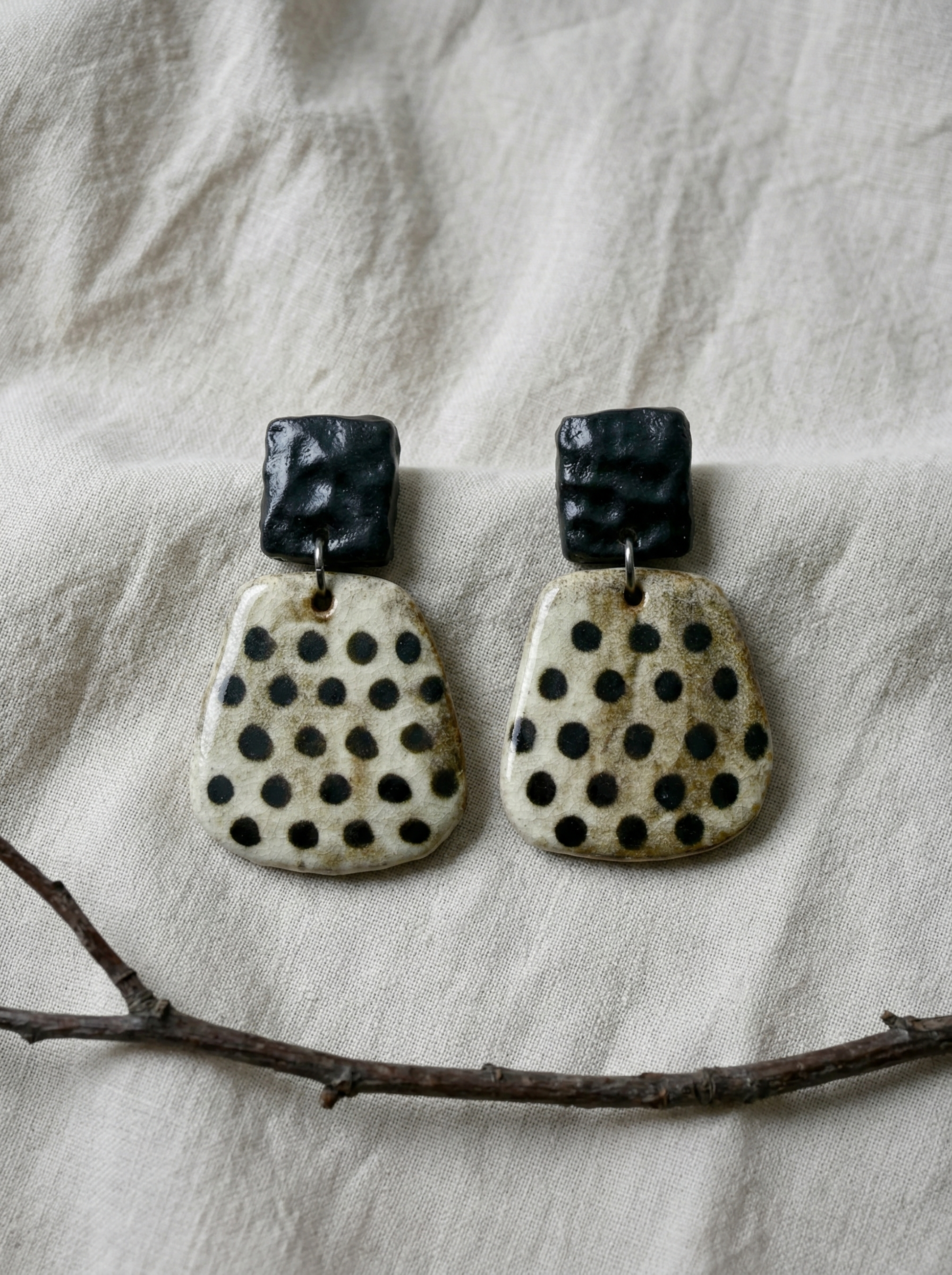 A pair of HECROWN wabi-sabi ceramic earrings with black hammered tops and speckled dot bottoms on natural linen. Unique handmade jewelry from Jingdezhen.